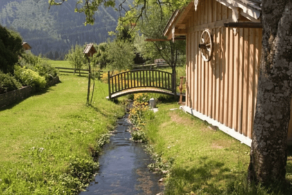 Kleiner Bach zwischen 2 Wiesen. Holzbrücke führt hinüber, an der rechten Seite zum Haus